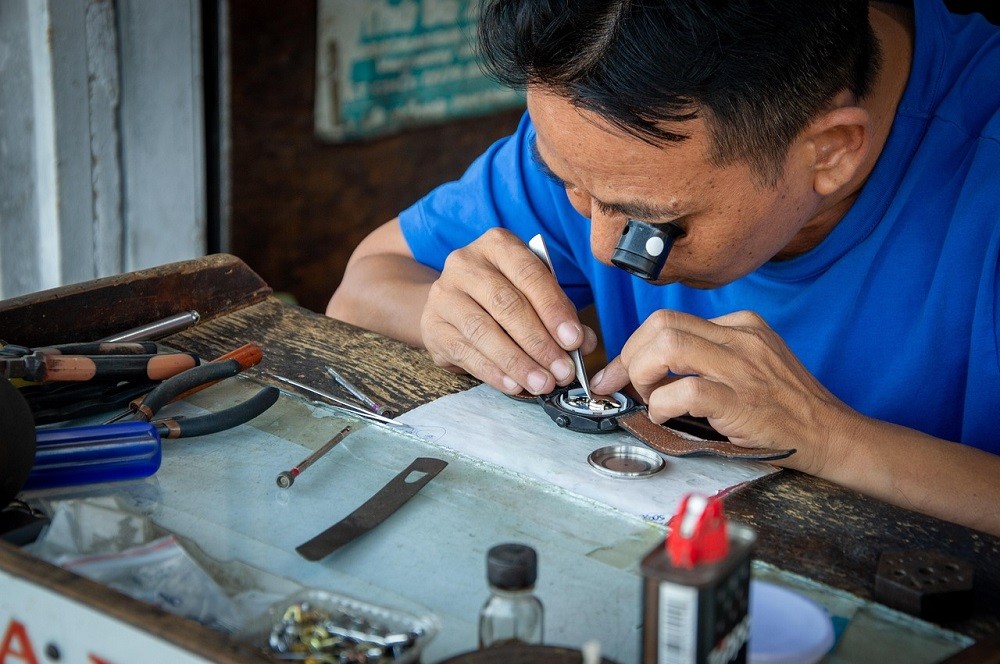 A man is repairing a watch. He has the back cover removed and he is loosening a screw on the movement