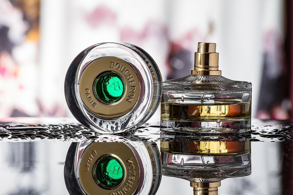 A short, round bottle of cologne has a brass atomizer. The clear glass cap is sitting upright beside the bottle