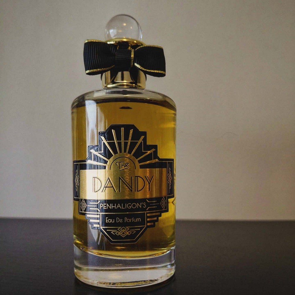 A bottle of The Dandy by Penhaligons contains light brown liquid and has a black and gold art-deco inspired sticker with the brand and fragrance information on it