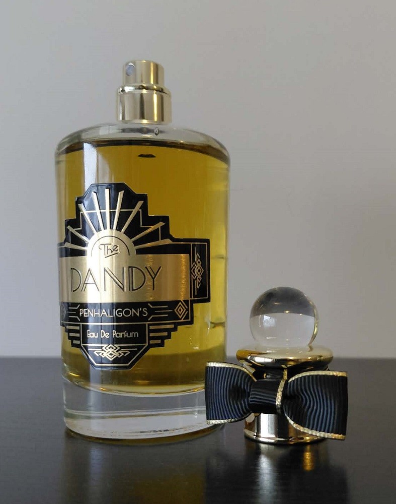 A light brown bottle of The Dandy cologne by Penhaligon's has its cap with a miniature black bow tie sitting beside it
