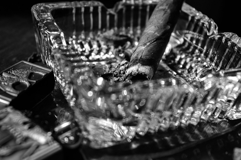 A black and white photograph of a smoked cigar sitting in a square glass ashtray