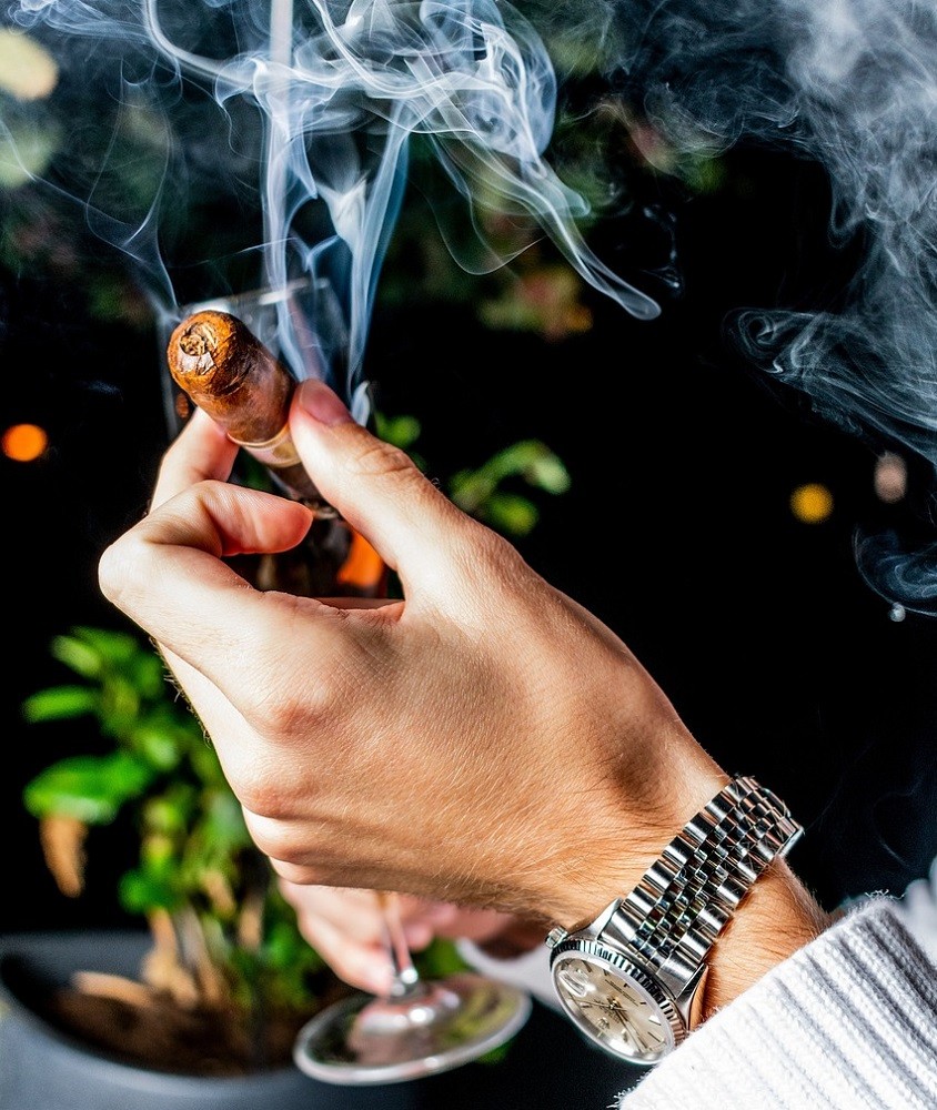 A man is holding a light brown cigar. Silky white smoke is coming out of the lit end of the cigar