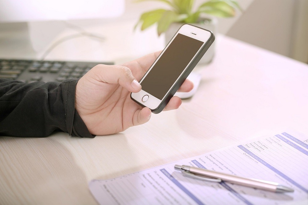 A man is holding out his cell phone and resting his hand on his desk