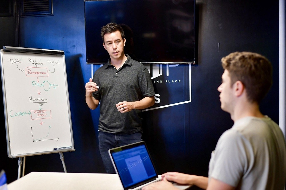 A young man points to a whiteboard while someone seated nearby is watching him