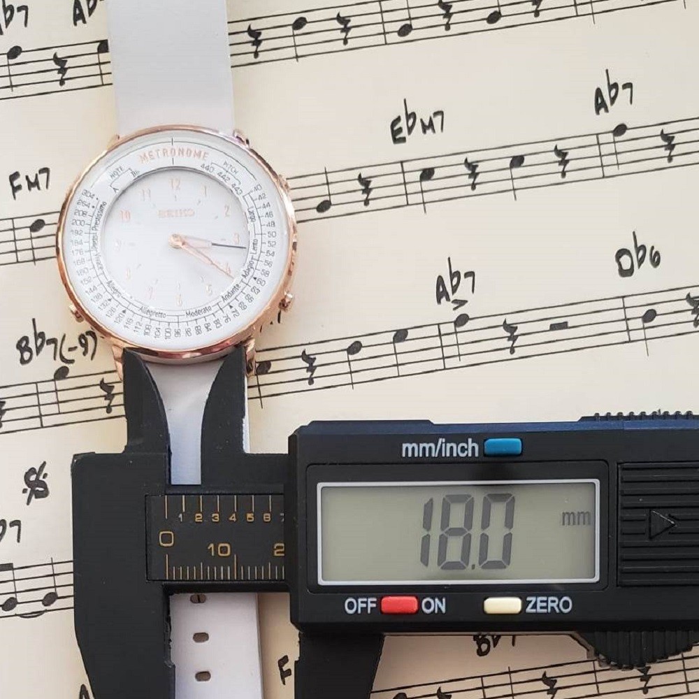 A white Seiko Metronome watch is measured with calipers. The measurement reads 18mm, the width of the watch strap