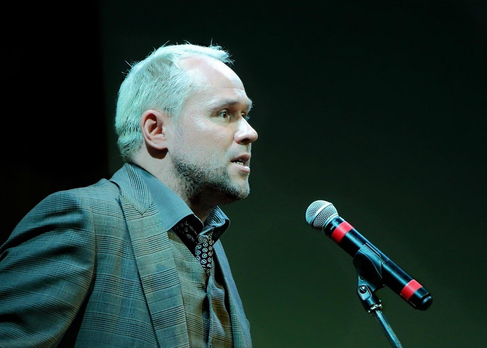 A man in a suit stands in front of a microphone
