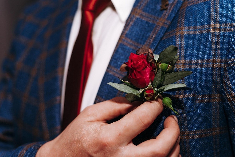 A man wearing a blue suit is adjusting his red rose boutonniere