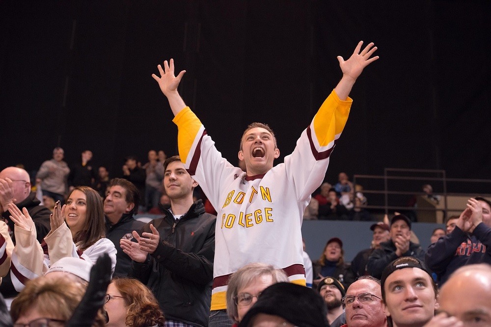 A fan wearing a white and yellow jersey is standing up and cheering