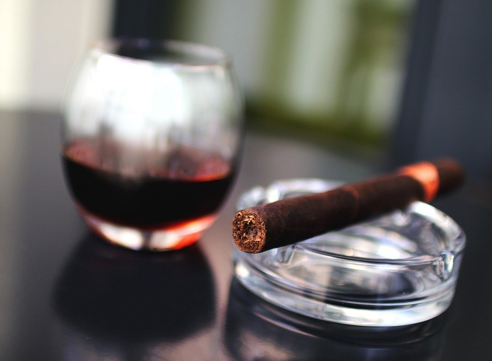 A cigar sits on a glass ashtray and a glass of bourbon is behind it