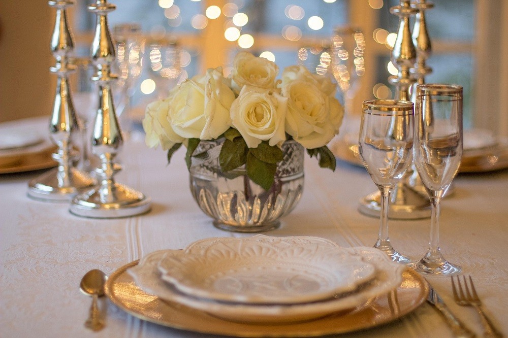 A formal place setting shows decorative forks, knives, spoons, plates, glasses, and a small bouquet of white roses on the table