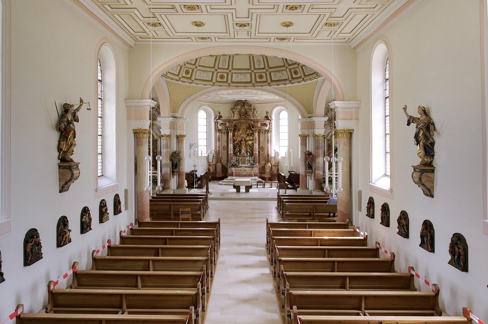 Interior view of a church with white walls, brown pews, and bronze angel decorations on the walls