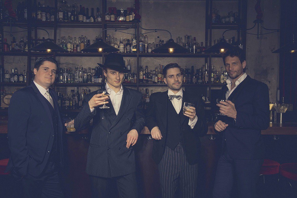 Several gentlemen are at a bar holding glasses to toast the viewer