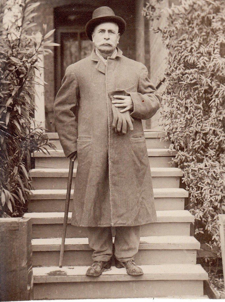 A black and white photograph shows an older man in a dress coat holding gloves and a walking cane