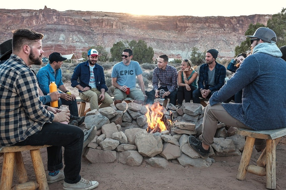 A group of friends sit around a lit campfire. They are all smiling and laughing