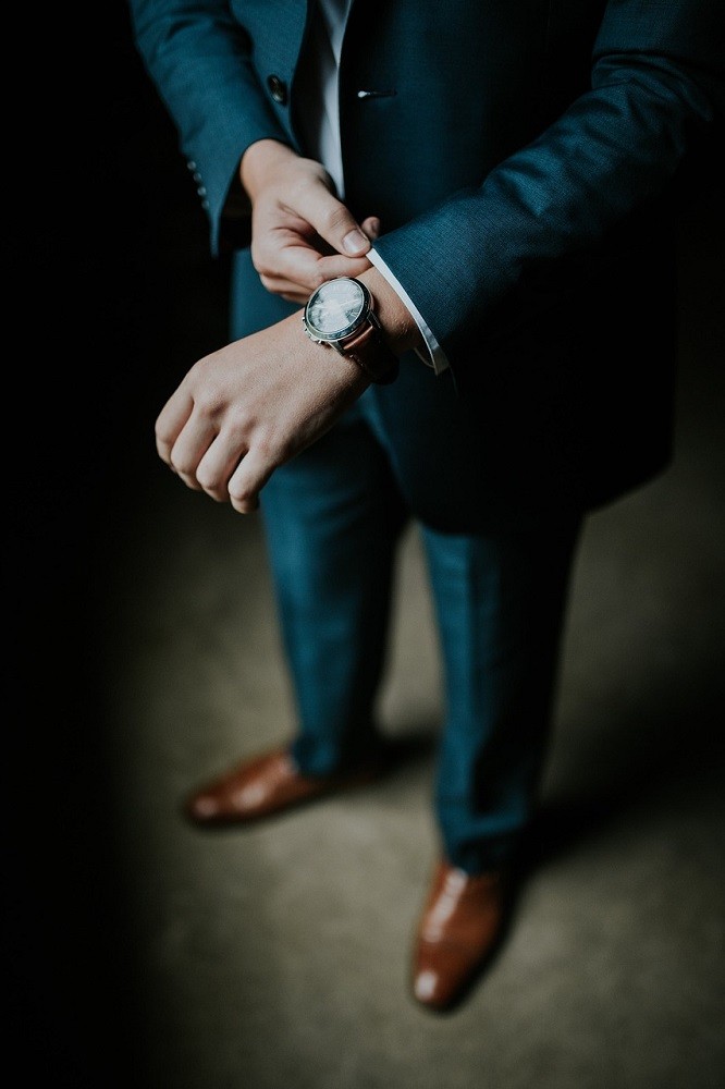A well-dressed man in a blue suit and brown dress shoes is adjusting his Rolex watch