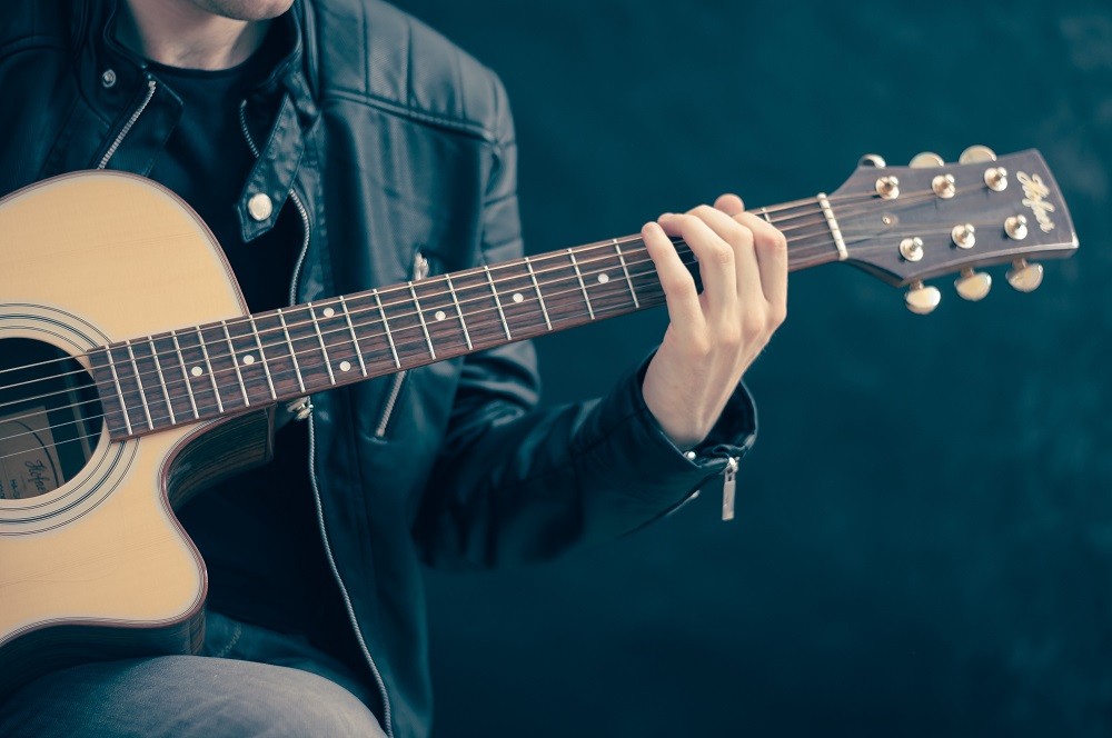 A young man in a leather jacket is playing an acoustic guitar on a bench