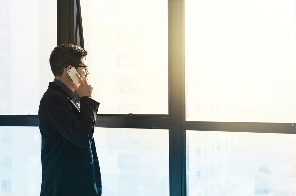 A businessman looks out his window. He may be in a meeting but takes his call outside. He's wear a black suit, has short black hair, and holding a white smartphone