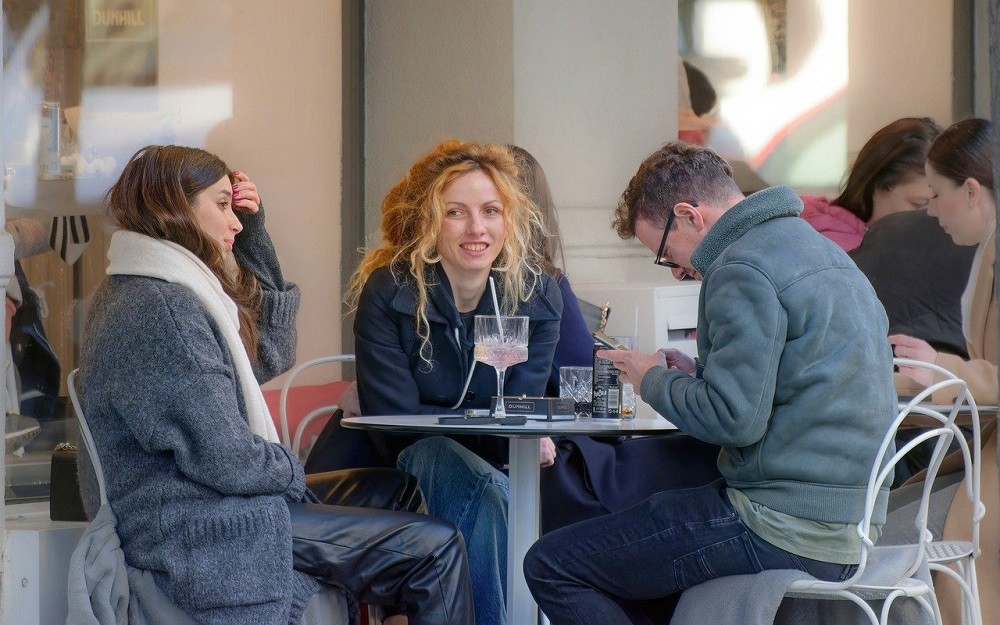Three people, a man and two women, sit at a table for a meal. The man is looking down at his phone, phubbing the two women. The two women look annoyed
