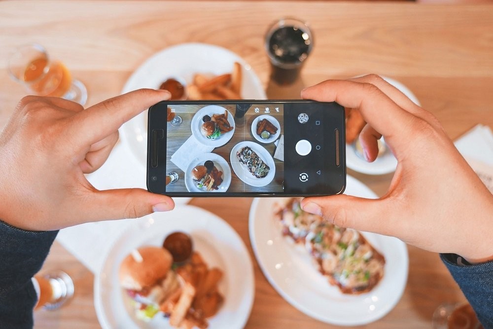 A phone is held up high above plates of food. A person is taking a picture of all the dishes in front of them