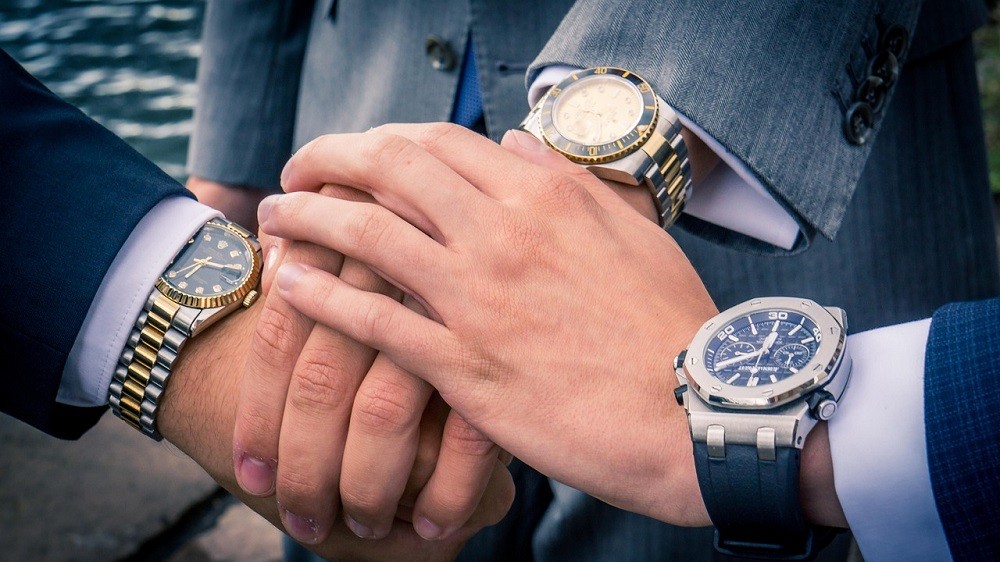 Three businessmen are shaking hands. They are all wearing expensive luxury watches like Rolex, Tudor, and Audemars Piguet