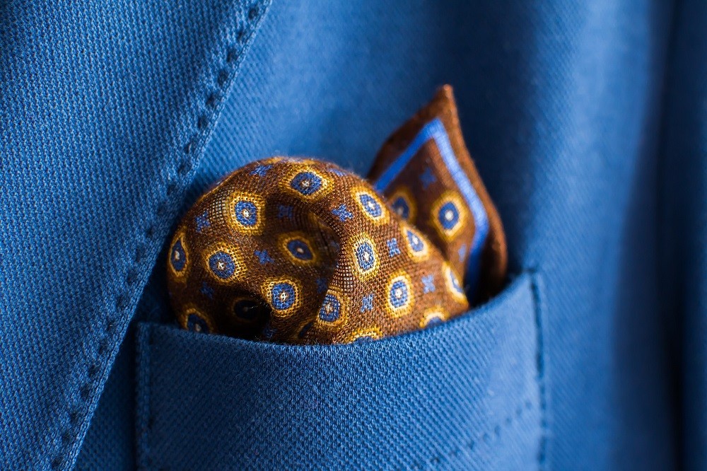 A brown pocket square with blue and yellow dots sits inside the breast pocket of a lighter blue suit