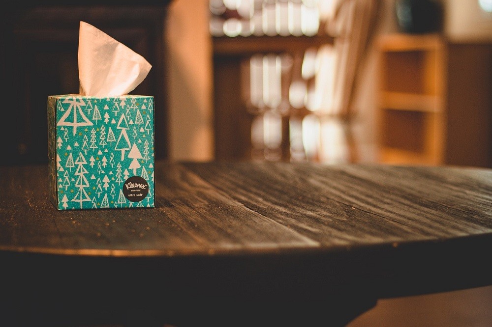 A square box of Kleenex brand tissue paper sits on a dark brown wooden table